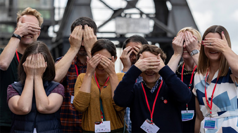 A group of people standing together, covering their eyes and smiling.