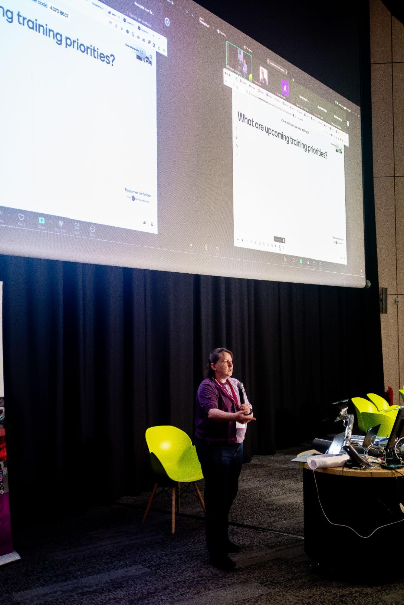 Samantha Ahern standing at a lectern, giving a presentation. The slide on the screens behind her says "what are upcoming training priorities?"