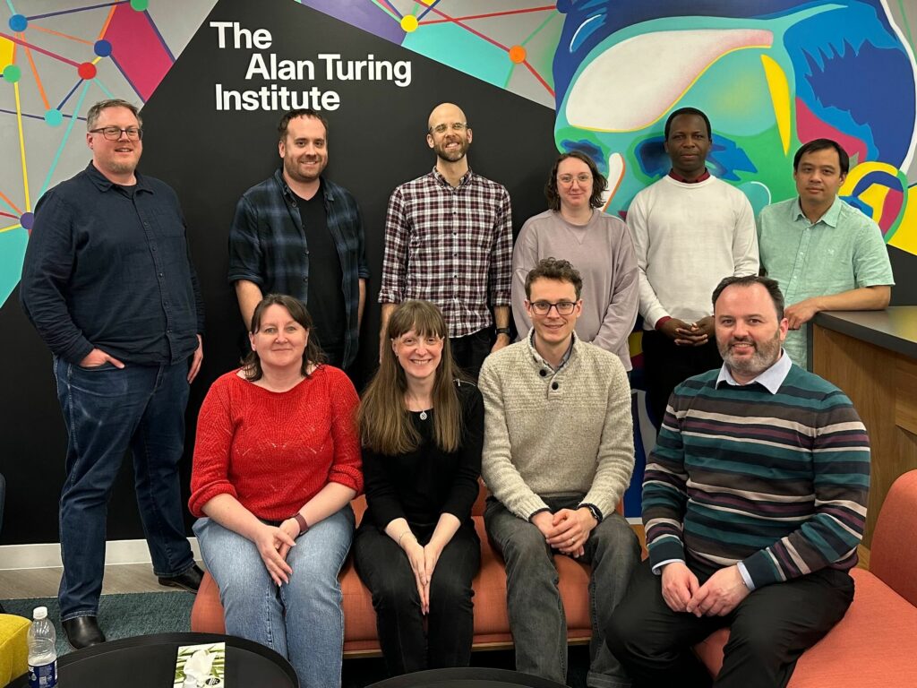 A photo of the trustees who attended an in-person meeting in March 2025. On the back row is David Beavan, Mike Simpson Stef Piatek, Fliss Guest, Godwin Yeboah and Twin Karmakharm. On the front row: Samantha Ahern, Gillian Sinclair, Pip Grylls and Martin O'Reilly. They are standing in front of the mural of Alan Turing at the Alan Turing Institute in London.