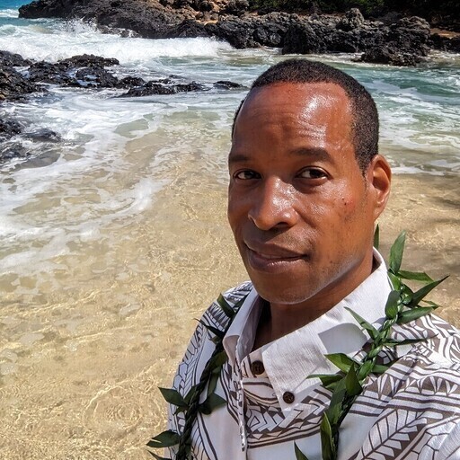 Photo of Darold Davis standing on a beach, wearing a patterned shirt and a green leafy lei. 
