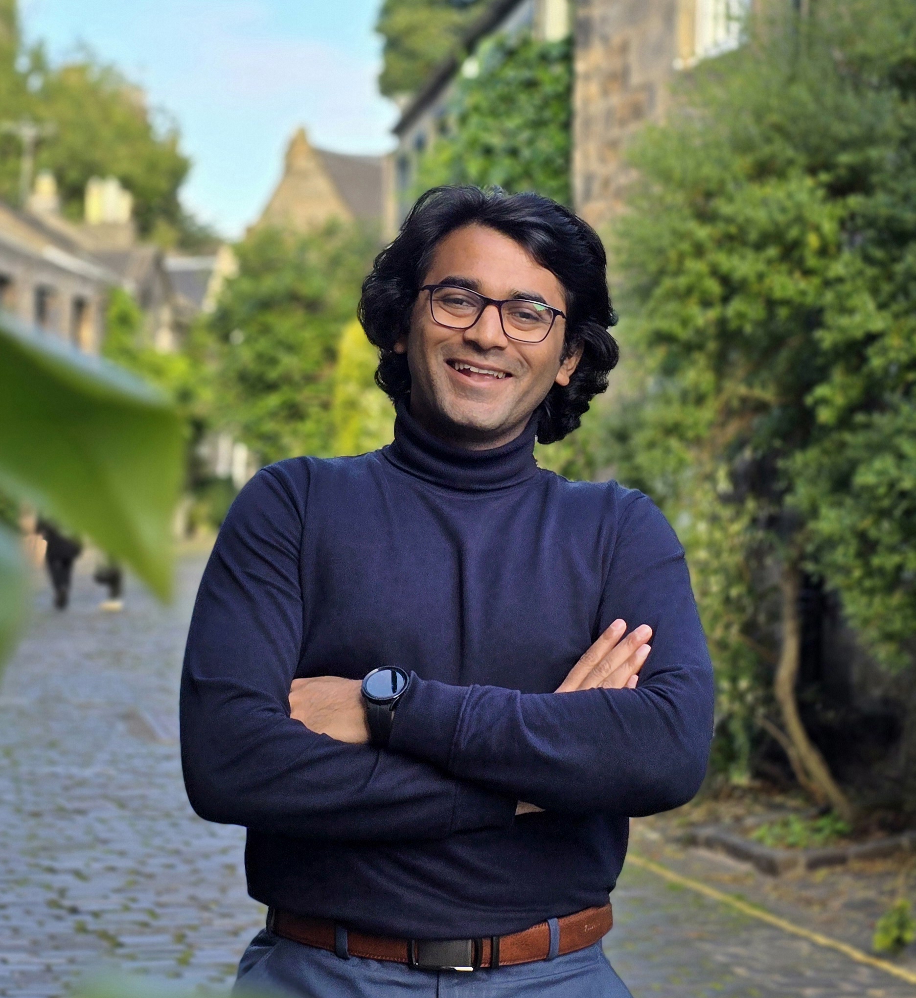 Photo of Prasad Dilip Sutar standing on a verdant, cobbled street. He's wearing a black turtleneck and glasses.