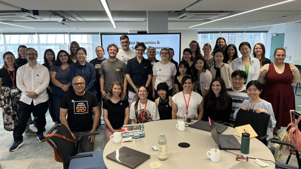 A photo of around 30 people, smiling at the camera. Behind them is a screen with a slide saying "Digital Humanities & Research Software Engineering Summer School 2025"