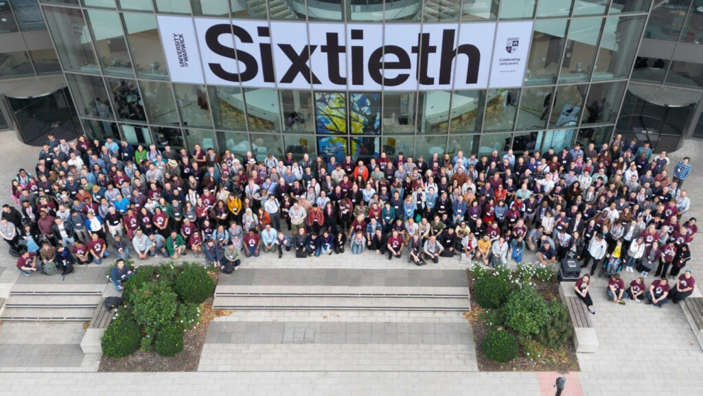A drone photo of the RSECon attendees. Around 400 people, standing in rows and looking up at the camera outside the Oculus building at the University of Warwick.