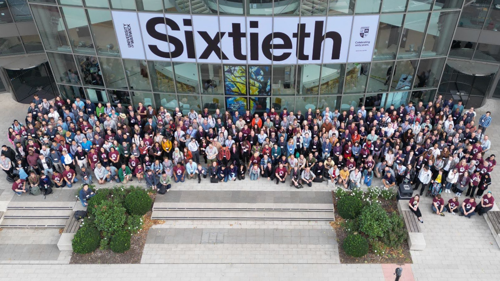 The group photo from RSECon25. A drone shot of 400 people standing together in front of the Oculus building at the University of Warwick.